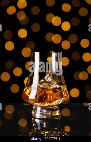 Whiskey in einem Tulpenglas mit Eiswürfeln, das goldenes Licht vor einem dunklen Hintergrund mit orangefarbenem Bokeh reflektiert Stockfoto