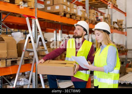 Männlicher Mitarbeiter mit Box, der in die Lagerregale schaut, während eine Kollegin die Dokumentation eingeht. Strategische Planung und Lagerorganisation im Vertriebszentrum Stockfoto