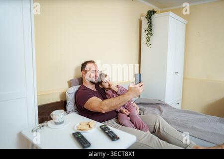 Glücklicher Vater und Tochter, die sich im Bett entspannen und ein Selfie mit einem Smartphone machen Stockfoto