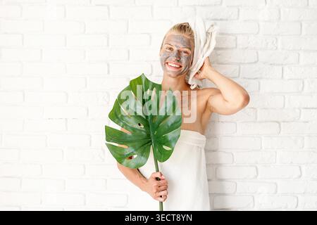 Spa Gesichtsmaske. Spa und Schönheit. Fröhliche lächelnde Frau mit Badetüchern und einer Gesichtsmaske aus Ton auf ihrem Gesicht, die ein grünes Monstera-Blatt hält Stockfoto