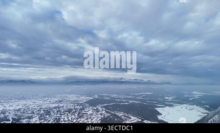 Breites Luftpanorama einer Winterstadt und eines sich windenden Flusses unter hoch aufragenden Wolken und treibendem Nebel. Schneedecken, Dächer und Felder Stockfoto