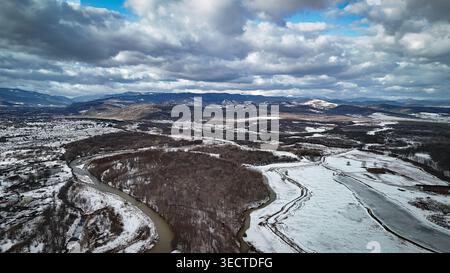 Die Luftperspektive zeigt ein winterliches Tal aus schlängelnden Flüssen, Wäldern und schneebedeckten Feldern, die von dunklen Bergrücken eingerahmt werden. Schwere Wolken ziehen über sich Stockfoto