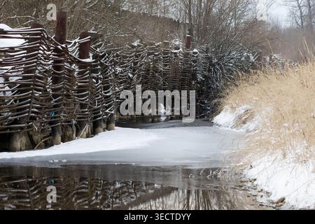 Winterlandschaft mit einem schmalen gefrorenen Fluss neben einem rustikalen gewebten Holzzaun und trockenem Gras. Weicher Schnee bedeckt den Boden und schafft eine ruhige ländliche Szene Stockfoto