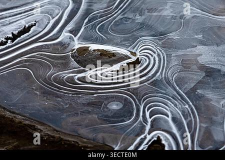 Ein grafisches Muster aus eisigen Ringen und Rillen umgibt eine dunkle Öffnung im gefrorenen Fluss. Helle Grate zeichnen energetische Pfade auf Stockfoto
