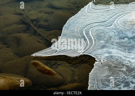 Eine dünne dreieckige Eisflosse erstreckt sich über flaches Wasser wie ein Glassegel. Konzentrische Bänder bogen zu einer scharfen Spitze, während glatte Steine darunter liegen. Stockfoto