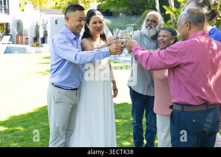 Braut in weißem Kleid und Schleier hebt Glas und initiiert Hochzeitstoast, Gäste klirren auf dem Rasen Stockfoto
