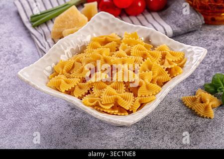 Lebendiges Stillleben. Trocknen Sie Farfalle Nudeln in einer weißen Schüssel. Frische Kirschtomaten, Parmesankäse, Basilikumblätter, grüne Zwiebeln, Flasche Olivenöl Stockfoto