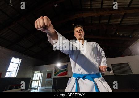 Ein fokussierter Karate-Schüler mit weißem Gi und blauem Gürtel sorgt in einer geräumigen Trainingshalle für einen starken Schlag. Dramatische Beleuchtung mit niedrigem Winkel und einem breiten Stockfoto
