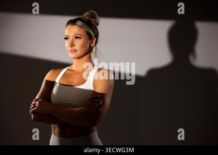 Selbstbewusste junge Frau in weißem Sport-BH mit überkreuzten Armen in dramatischer Beleuchtung Stockfoto