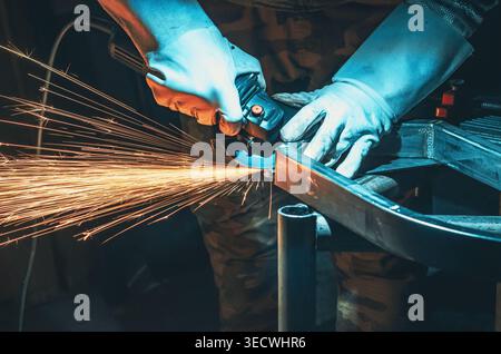 Handwerker in Schutzhandschuhen mit Schleifmaschine auf dunklem Hintergrund. Goldene Funken beim Schleifen von Metall in der Werkstatt. Metallbearbeitung. Stockfoto