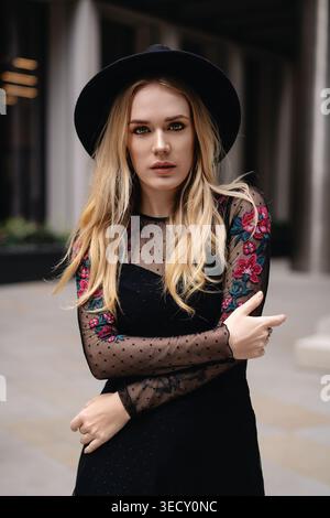Die junge blonde Frau posiert in einer modernen Stadt für ein Outdoorporträt mit einem schwarzen Hut mit breiter Krempe und einem schwarzen Kleid mit durchsichtigen Ärmeln Stockfoto