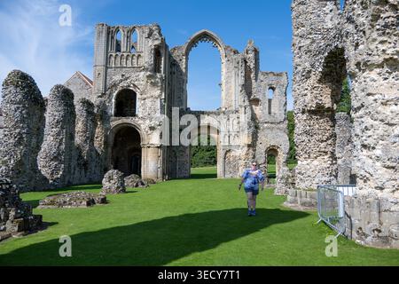 Ein Besucher, der auf dem Rasen vor der verfallenen Kirchenfassade und den Bögen in Castle Acre Priory, Castle Acre, Norfolk, England, Großbritannien spaziert. Stockfoto