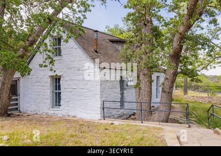 Ein bezauberndes weißes Steinhaus mit einem Holzdach steht eingebettet zwischen hohen Laubbäumen neben einem gepflasterten Gang. Stockfoto