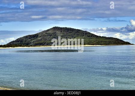 Ein üppiger, grüner Hügel erhebt sich von einer Sandküste und trifft im Vordergrund auf ruhiges, lebendiges blaues Meereswasser. Weiche Wolken ziehen über einen hellen Himmel und fügen hinzu Stockfoto