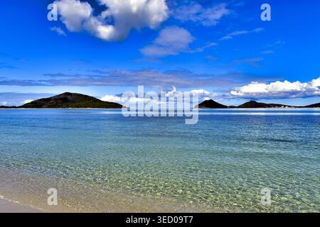 Klares, flaches türkisfarbenes Wasser offenbart den sandigen Meeresboden, bevor es sich zu einer Linie kleiner, grüner, hügeliger Inseln unter einem hellblauen Himmel vertieft. Weich mit Stockfoto
