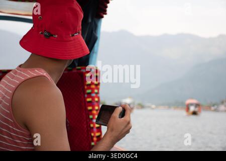 Ein Mann mit rotem Eimerhut und gestreiftem Tanktop macht ein Foto vom malerischen Dal-See in Srinagar, Kaschmir, während er auf einem traditionellen Schiff fährt Stockfoto