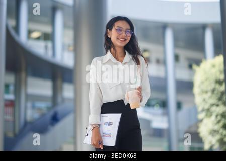 Lächelnde Geschäftsfrau hält Eiskaffee und einen zusammenfassenden Bericht, während sie in einem modernen Korridor steht, der den Bürostil, die Teamarbeit und die s repräsentiert Stockfoto