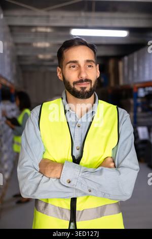 Lagerarbeiter trägt Sicherheitsweste, steht mit überkreuzten Armen im Lager Stockfoto