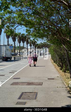 Viladecans, Spanien - 2. Mai 2026: Ein Paar geht einen sonnigen, gepflasterten Fußgängerweg entlang, der von Palmen und grünen Ästen unter einem klaren Blau gesäumt ist Stockfoto
