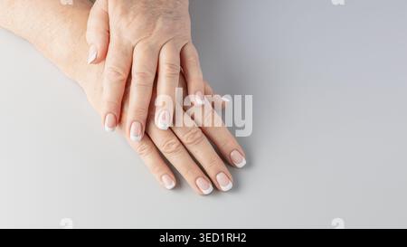 Französische Nagelmaniküre an alternden Händen einer erwachsenen 65-jährigen Frau mit Altersflecken, tiefen Hautfalten. Hautalterung und Nagelpflegekonzept im Alter. Weiblich Stockfoto