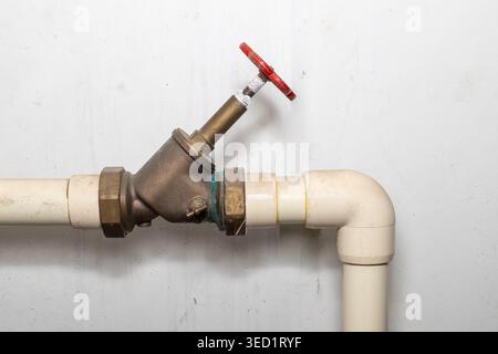 Detail des staubigen Wasserrohrsystems mit roter Armatur im Untergeschoss des Wohnhauses. Staubige Rohrleitungen auf schmutzigem weißem Wandhintergrund mit Kopierraum. Stockfoto