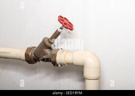 Detail des staubigen Wasserrohrsystems mit roter Armatur im Untergeschoss des Wohnhauses. Staubige Rohrleitungen auf schmutzigem weißem Wandhintergrund mit Kopierraum Stockfoto