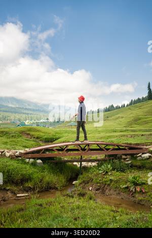 Ein Mann mit rotem Eimerhut, der über eine rustikale Holzbrücke über einen kleinen Bach in einer malerischen, grasbewachsenen Berglandschaft unter blauem Himmel spaziert. Stockfoto