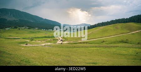Ein atemberaubender Panoramablick auf eine riesige grüne Wiese in einem Bergtal mit sanften Hügeln, einem unbefestigten Pfad und einem dramatischen bewölkten Himmel mit Sonnenschein Stockfoto