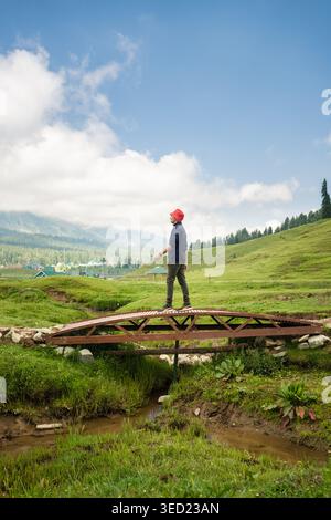 Ein Mann mit rotem Eimerhut, der über eine rustikale Holzbrücke über einen kleinen Bach in einer malerischen, grasbewachsenen Berglandschaft unter blauem Himmel spaziert. Stockfoto