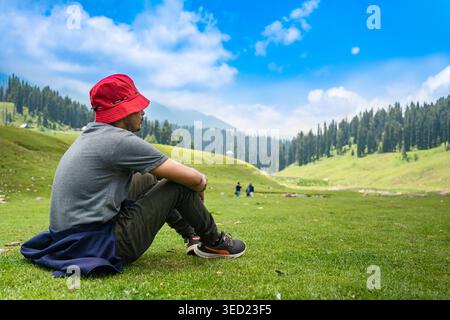 Ein Mann mit rotem Hut sitzt auf dem üppigen grünen Gras eines Bergtals in Kaschmir, Indien, und genießt die malerische Aussicht auf die umliegenden Kiefernwälder Stockfoto