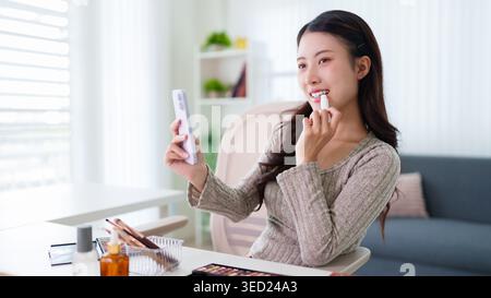 Junge asiatische Frau, die Lippenstift anwendet und auf den Smartphone-Spiegel schminkt, die Schönheitsroutine zu Hause im Wohnzimmer macht Stockfoto