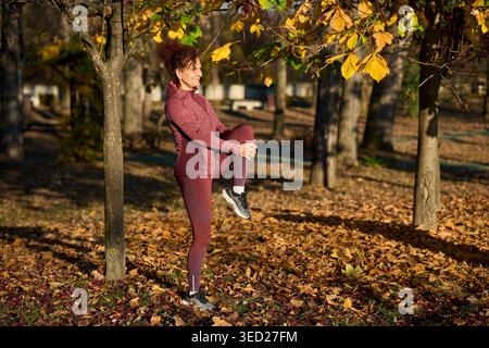 Eine Frau mittleren Alters in burgunderroter Sportbekleidung, die auf einem Bein steht und ihr hochgezogenes Knie dehnt und sich vor einem Lauf durch die goldene herbstlea aufwärmt Stockfoto