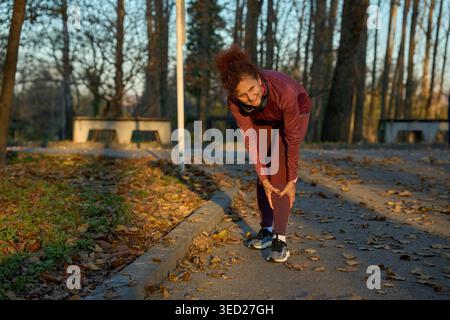 Eine Frau mittleren Alters in burgunderroter Sportbekleidung beugte sich und grimmierte, während sie ihr Schienbein auf einem Parkweg festhielt, was auf Schmerzen durch eine Laufverletzung wie eine deutet Stockfoto