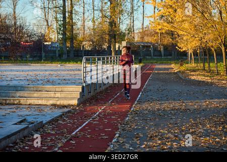Frau mittleren Alters in burgunderroter Sportbekleidung, die auf einer roten Laufstrecke neben Stadiontribünen in einem sonnigen Herbstpark joggen, umgeben von goldenem Laub Stockfoto