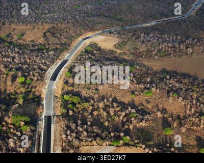 Eine Straße durchquert ein trockenes Gebiet mit kargen Bäumen und offenem Land am Nachmittag. Die Aussicht zeigt eine Mischung aus Schmutz und Bürgersteig. Stockfoto
