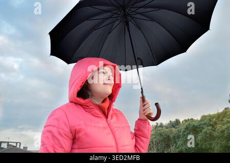 Schöne junge Frau, die einen rosafarbenen Anorak trägt, hält im Herbst einen schwarzen Regenschirm, überrascht und aufgeregt unter dem angehaltenen Regen Stockfoto