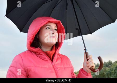 Die schöne junge Frau, die im Herbst einen rosafarbenen Anorak mit einem schwarzen Regenschirm trug, war überrascht und aufgeregt unter dem Regen Stockfoto