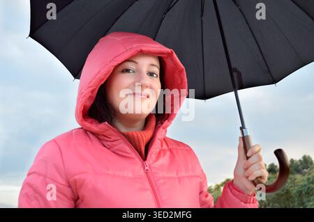 Eine schöne junge Frau, die einen rosa Anorak mit Kapuze trägt, hält einen schwarzen Regenschirm an einem herbstlichen Regentag auf dem Land Stockfoto