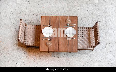 Aus der Vogelperspektive eines leeren Cafés. Ein quadratischer Holztisch und gewebte Lederstühle sind symmetrisch auf einem hellen, gesprenkelten Terrazzo-Boden platziert. Stockfoto