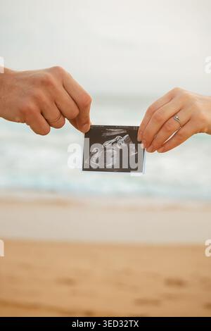 Das Paar hält Ultraschallfotos am Strand während Sonnenuntergang, während die Wellen im Hintergrund abstürzen. Zwei Menschen stehen und erscheinen glücklich und genießen den Moment als Su Stockfoto