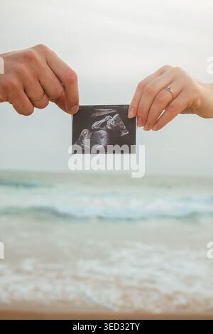 Das Paar hält Ultraschallfotos am Strand während Sonnenuntergang, während die Wellen im Hintergrund abstürzen. Zwei Menschen stehen und erscheinen glücklich und genießen den Moment als Su Stockfoto