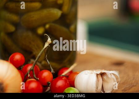 Frische Kirschtomaten und Knoblauch im Vordergrund mit einem Glas eingelegter Gurken im Hintergrund. Traditionelles Konservierungskonzept. Stockfoto