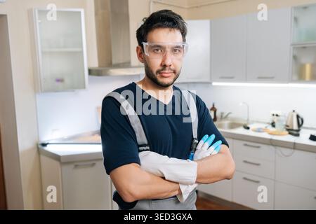 Professioneller männlicher Arbeiter mit Schutzbrille und Arbeitshandschuhen, überkreuzte Arme, während er selbstbewusst in der modernen Küche steht, bereit für die Reinigung Stockfoto