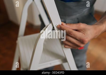 Nahaufnahme der Hand des Mannes, die die weiße Metall-Trittleiter öffnet. Arbeiter in grauen Overalls, die Geräte für die häusliche Verbesserung oder Reparatur vorbereiten. Geringe Tiefe von Stockfoto