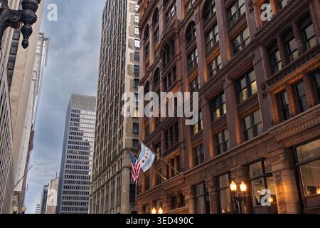 Die Leute laufen entlang der Straße in der Innenstadt von Chicago, während die Gebäude auf beiden Seiten hoch stehen. Flaggen winken sanft im Wind gegen einen bewölkten Himmel während der Th Stockfoto