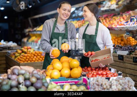 Zwei positive weibliche Vertriebsmitarbeiter stellen frisches Gemüse und Obst in die Regale des Supermarktes Stockfoto