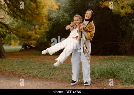 Glückliche Mutter hebt ihre Tochter im Herbst in einem Park und genießt einen verspielten Moment zusammen. Familienkonzept mit Liebe, Freude und Verbundenheit Stockfoto