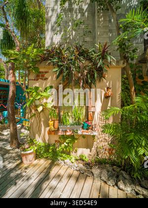 USA, Islamorada, FL - 18. April 2026: Ein dekoratives Kunstfenster ist in eine Außenwand mit freiliegenden roten Ziegeln und tropischen Pflanzen eingebaut. Das künstlerische Stockfoto
