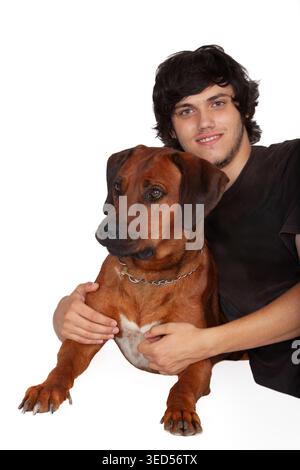 Ein junger Mann mit dunklen Haaren in einem schwarzen T-Shirt ringt spielerisch mit einem braunen Rhodesian Ridgeback-Hund, der auf dem Rücken liegt. Stockfoto