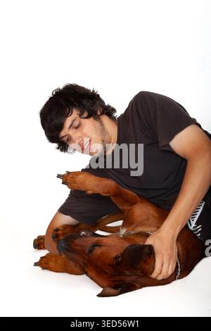 Ein junger Mann mit dunklen Haaren in einem schwarzen T-Shirt ringt spielerisch mit einem braunen Rhodesian Ridgeback-Hund, der auf dem Rücken liegt. Stockfoto
